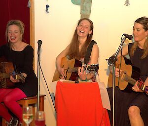 Three expeditioners sit holding guitars with broad smiles on their faces as they play music and sing songs.