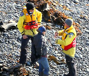 Tim and Paul awaiting pick up by the tourist boat tender in yellow wet weather gear on beach, with Jacque C