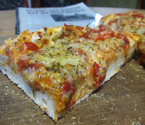 A thick slice of home made pizza sits on a wooden cutting board.