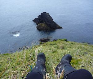 Man dangles boot clad feet down a hill. His perspective shows mostly water, with a large rock protruding from it.