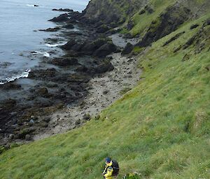 Female expeditioner with pack on the slopes of North Head, coast and water seen below