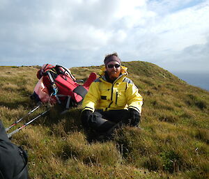 Male expeditioner with beard sits on grassy hill and looks toward camera