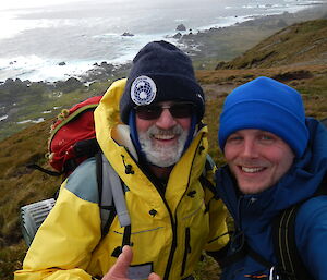 Two mail expeditioners smile for the camera and give a thumbs up. The image is a selfie style from a height so that the ocean and coast are visible below and behind the subjects.