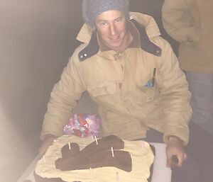 Male expeditioner in tradesman clothing holds up cake towards camera which features two short underwater hockey sticks and a puck made of icing.