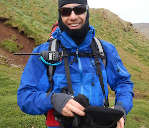 A male expeditioner on a grassy hill with only a third of his face visible. He is wearing cold weather clothes, and a walkie talkie is seen clipped to his shirt.