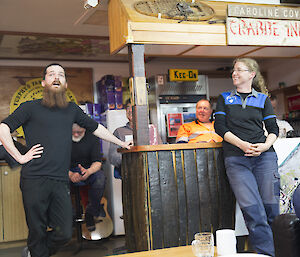 A bearded man speaking in front of a bar flanked by a woman, speaks to the crowd