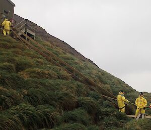 A wooden hut sits atop a small but steep hill covered in grass. One man at the top runs a rope down to a few people at the bottom.