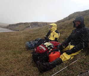 Two people sitting in all the wet weather gear in the lee of a hill side trying to stay out of the weather