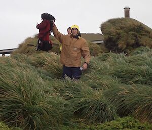 A man holds up the bag that represents a missing person in a tussock patch — the pack has a wig on top