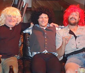 Three men on the couch wearing different coloured wigs