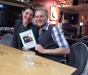 A man and a woman sit at a table holding a card that reads ‘Congratulations Marcus and Kim'