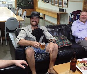 A man sits on the couch with a hat on and wig on his chest.