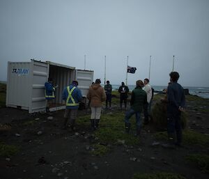 A group of people gather for the service on the beach with flagpoles in the background, on a very dark, wet weather day