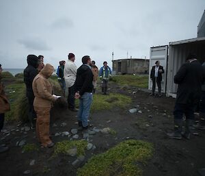 A man is reciting ANZAC poem ‘Ode to the Fallen’ to a group on a rocky beach in front of a small shipping container