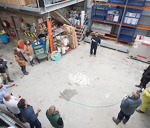 Taken from above inside a warehouse, this image depicts a group of people standing in a circle around a man flipping a coin