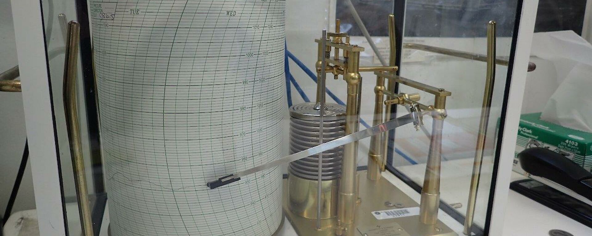 A beautiful old brass and glass barometer still working. It is inside a glass case, creating a graph on paper.