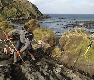 A man is digging in the dirt, with a muddy beach and water behind