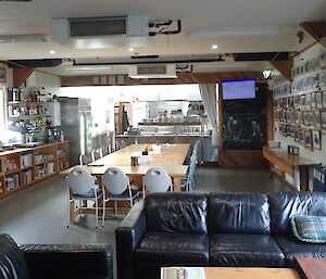 A shot of the mess looking towards the kitchen, with a large table through the centre, shelving for food on the left and a wall of photos on the right.