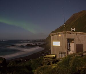The wooden hut that is Hurd Point with coast in background and an aurora in the sky