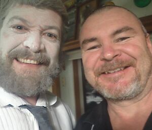 2 men smile at camera, one in white with talcum powder on face and one in back shirt.