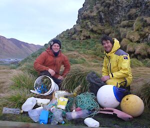 2 men in front of pile of debris they have collected on the beach which includes fishing buoys and lots of plastic