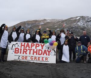 The team dressed as penguins around a happy birthday sign
