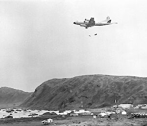 Black and white image of low flying aircraft dropping container with attached parachute. Station buildings and Wireless Hill in background