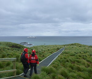 A ship off shore with some tourists on a boardwalk in foreground