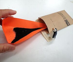 Putting seed into a paper bag using a funnel with ornamental penguin alongside
