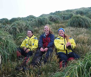 A trio of expeditioners sit smiling on the slope amidst tussocks of grass