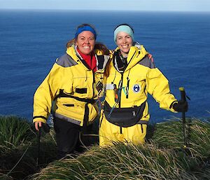 Two female albatross researchers standing on the top of a hill in green vegetation with the water behind them.