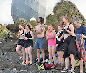 A group of expeditioners standing on a rocky beach in bathers looking cold.