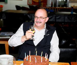 A man sitting at the table with a glass of wine in his hand and a cake sitting on the table in front of him.