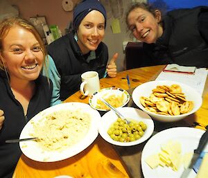 Three female expeditioners sit around a field hit table eating.