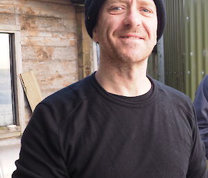 A man wearing a beanie standing outside of a wooden building.