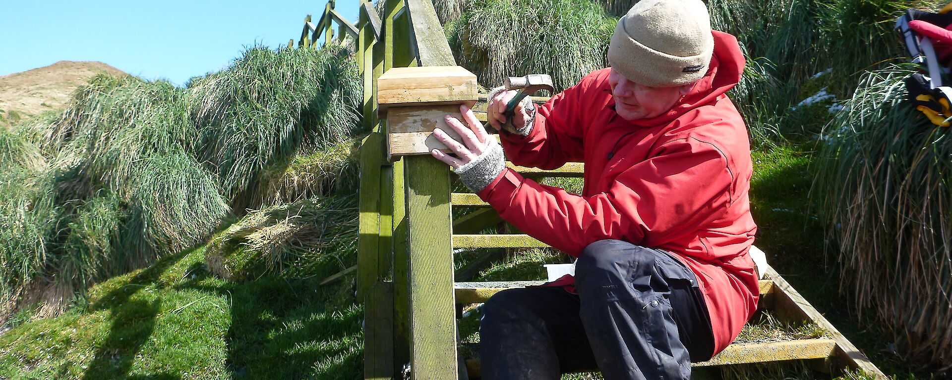 Man outdoors wearing a red raincoat and using tools to fix wooden staircase