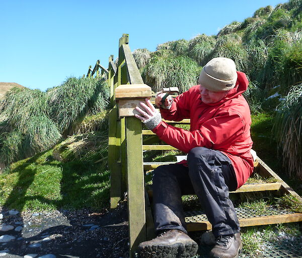 Man outdoors wearing a red raincoat and using tools to fix wooden staircase