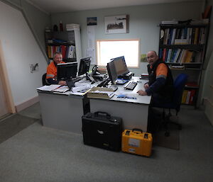 Two men sitting in an office at desks opposite each other and smiling at the camera