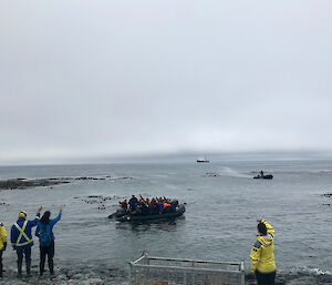People on the shore wave to people leaving in a small inflatable boat.