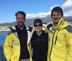 Three people standing on a ship in front of a mountain with a blue sky.