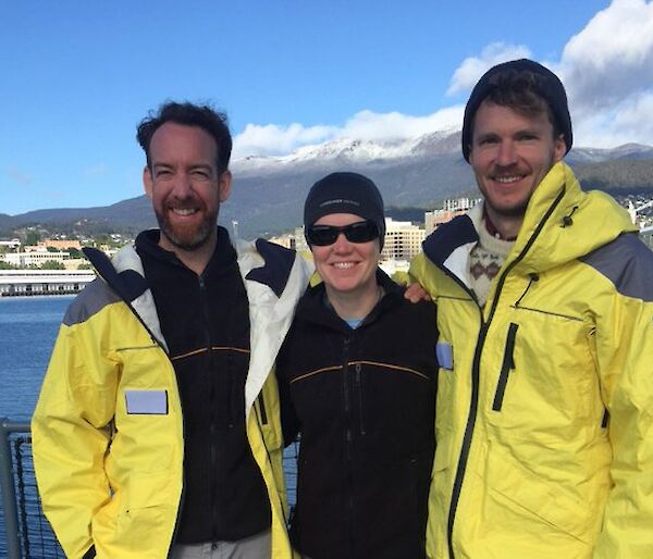 Three people standing on a ship in front of a mountain with a blue sky.