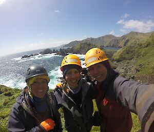 Three women wearing hard hats and outdoor jackets taking a selfie in front of a water landscape