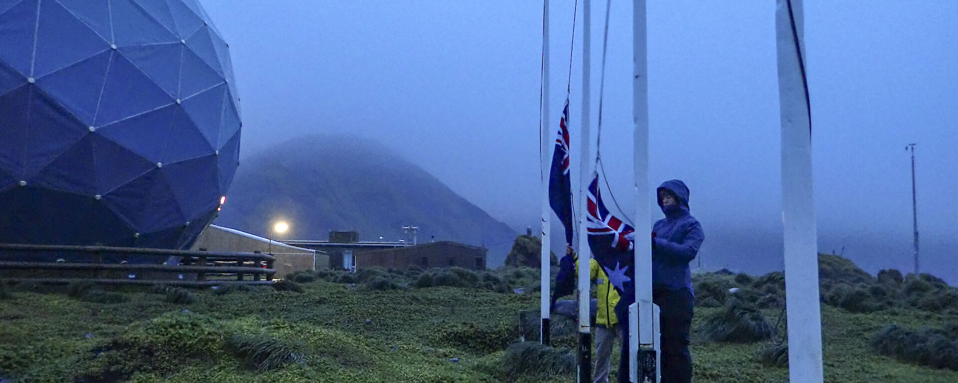 Two raincoat wearing epeditioners raise the NZ and Australian flags at dawn (0632) on the isthmus Macquarie Island
