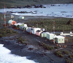 From Hut Hill looking south along the Isthmus in 1964 — Magnetic huts in background, from left to right — Nissen Hut — Mechanical workshop, radar, Nissen Hut — Meteorology store, balloon release, meteorological office, old wireless hut (14 sided Alaskan hut), old balloon release hut, communications office (stage1) and the meteorological screens in the fenced enclosure