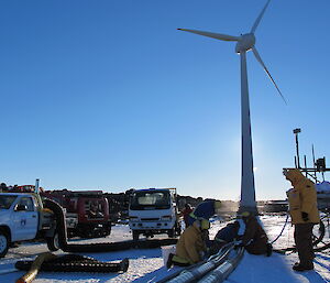 Three trucks and six expeditioners work on a large pipe with the Mawosn wind turbine towering in the background