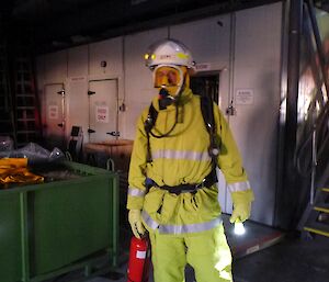 Male expeditioner in full fire fighting uniform, including breathing apparatus covering his face. He also holds a fire extinguisher