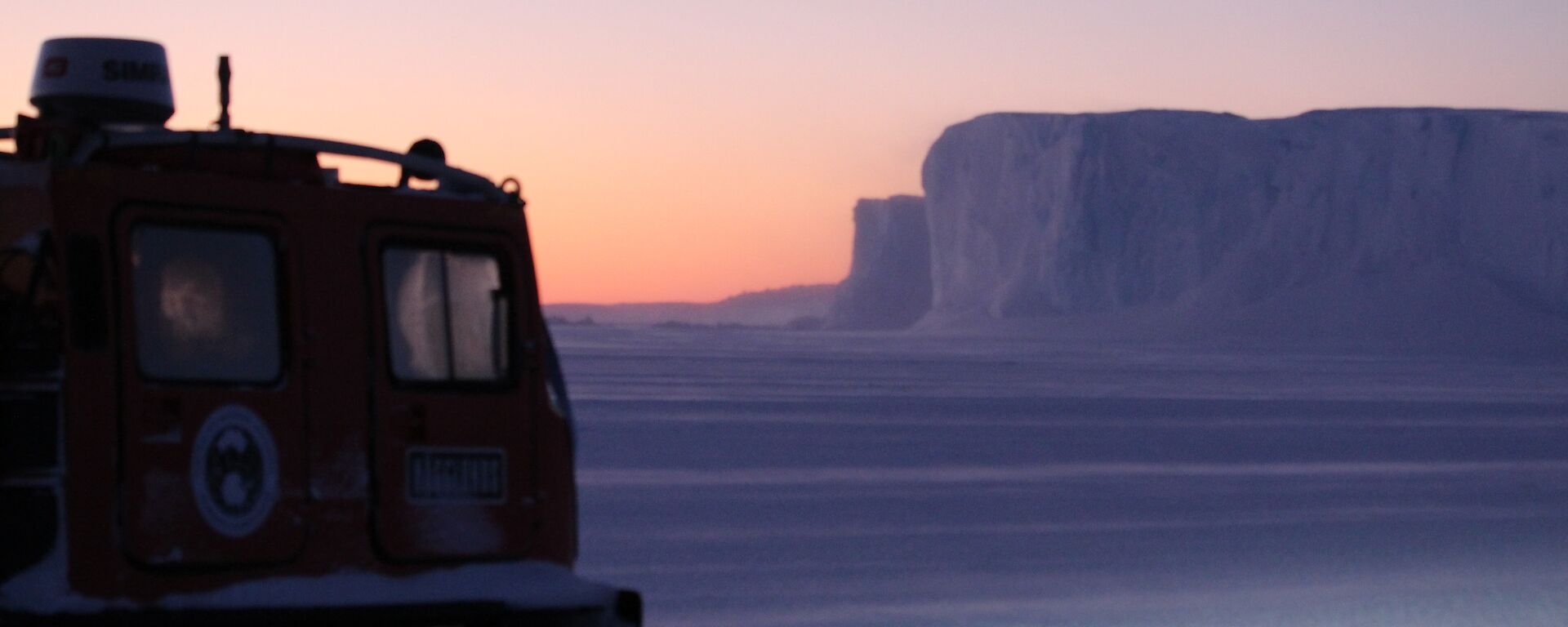 A Hägglunds with a iceberg in the background as the sun rises over the sea ice