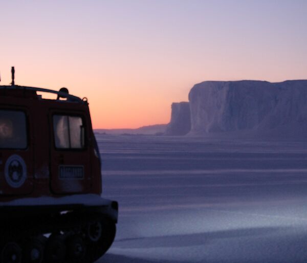 A Hägglunds with a iceberg in the background as the sun rises over the sea ice