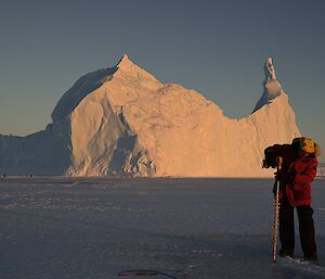 JB is drilling a hole to check the sea ice depth with an impressive iceberg in the background