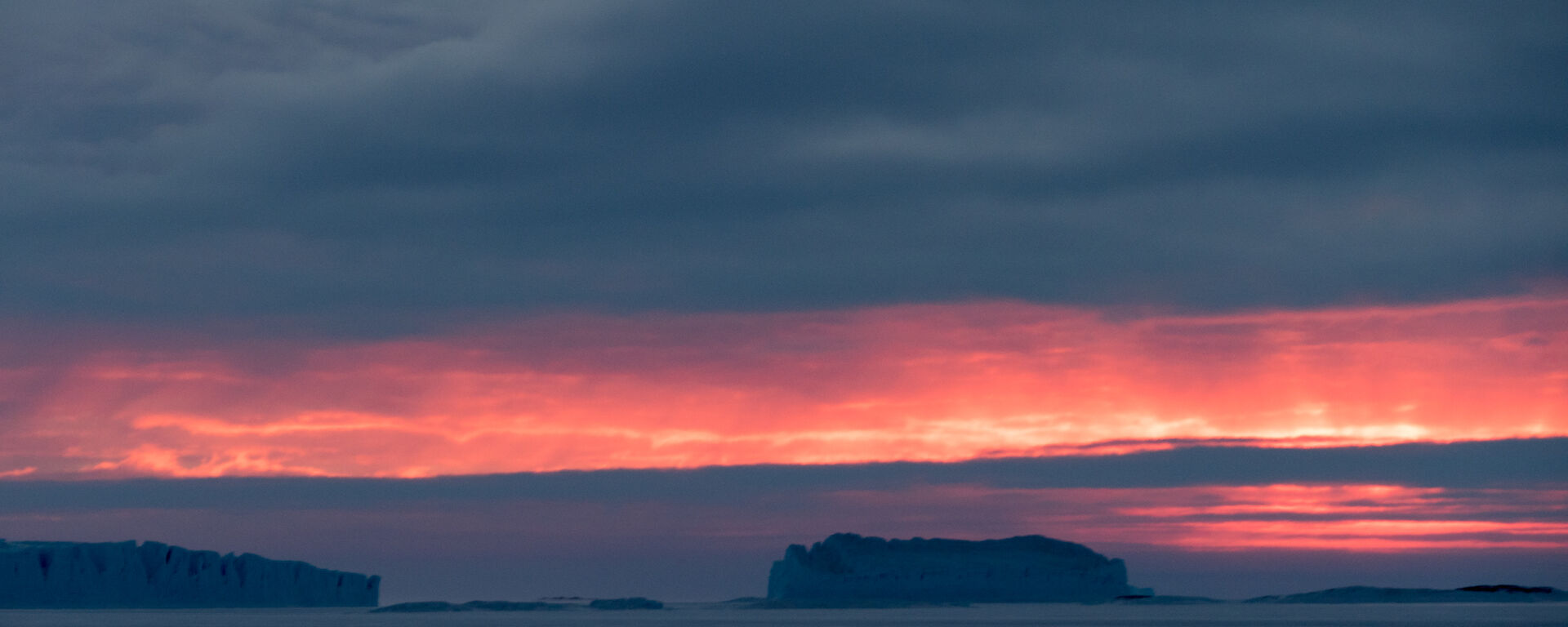 The horizon is glowing with orange to the north of Bechervaise Island over two massive icebergs